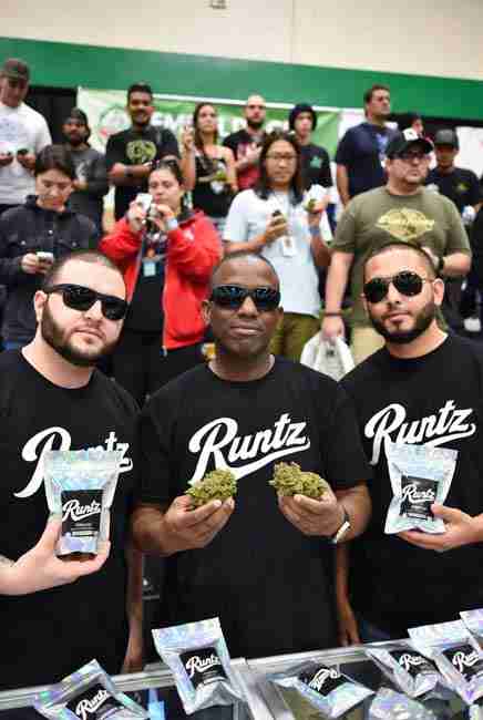 Runtz Cannabis breeders and team displaying colorful buds in iridescent mylar bags at cannabis cup event with crowd in background