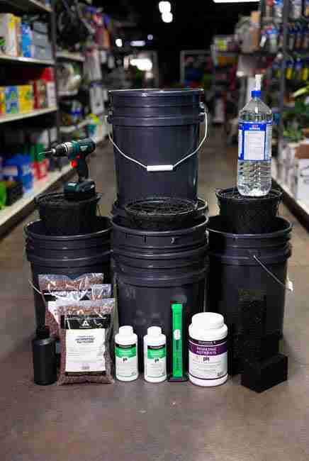 Complete collection of supplies for no pump hydroponics marijuana Kratky setup displayed in garden store setting with buckets, net pots, nutrients, measuring tools, and optional airstone for beginner Kratky cannabis setup 2026