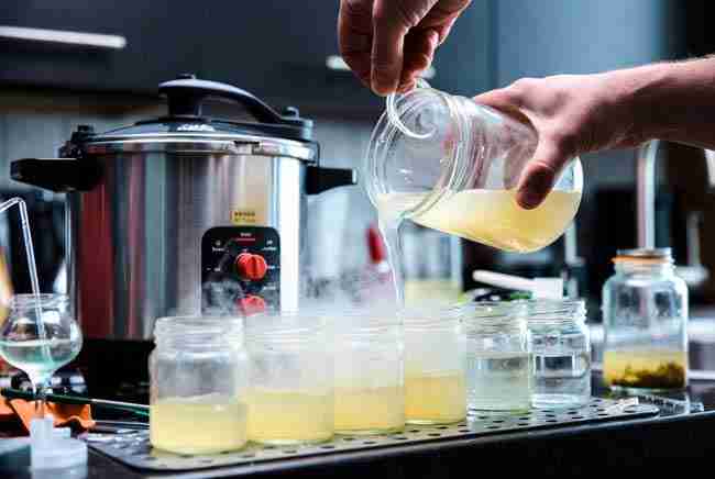 Pouring hot sterilized nutrient gel media into glass jars during tissue culture marijuana propagation setup with pressure cooker in background